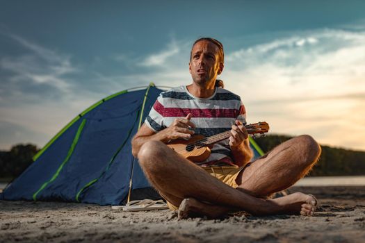 Happy young musician has a great time at the beach. He is sitting by the river, playing ukulele and singing. Sunset over water.