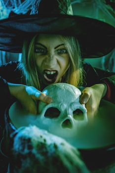 Close-up of a young witch with awfully face looking at camera and tells evil words. She is holds skull above cauldron with poison potion.