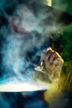 Close-up of young witch's hand with bone in creepy surroundings full of steam and fog, tells evil words to magic potion.