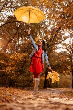 Happy young woman walking in autumn sunny park, holding umbrella height above her head and a branch of a fallen leaves in left hand.