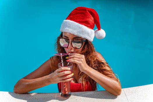 Young beautiful woman in the swimming pool in Santa Claus hat celebrating New Year and Christmas in hot country with glass of cocktail.