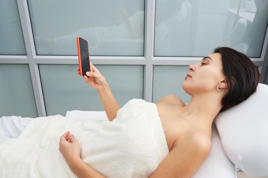 Attractive young woman using a smartphone and texting while laying on a massage bed at spa