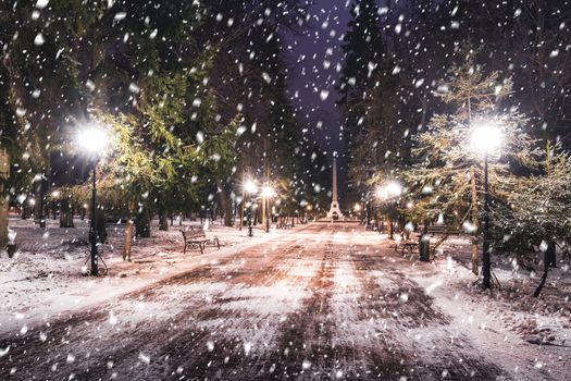 Snowfall in a winter park at night with christmas decorations, lights and  pavement covered with snow. Falling snow.