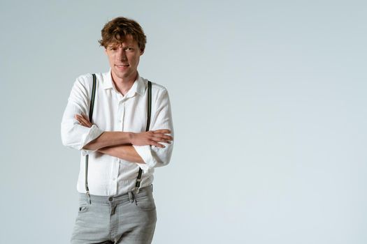 Handsome young man posing with hands folded dressed in white shirt and grey jeans isolated on white background. Copy space at right side.