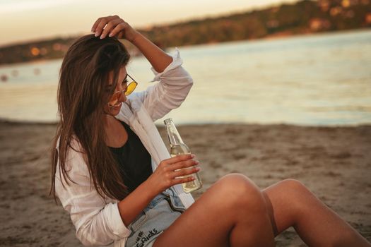 Young woman relaxing at sunset time on the river bank. She is sitting by the river and drinking beer. 