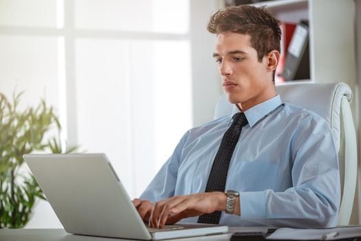 Executive male working at laptop looking at documents and accounts. He is typing and looking concentrated and serious.