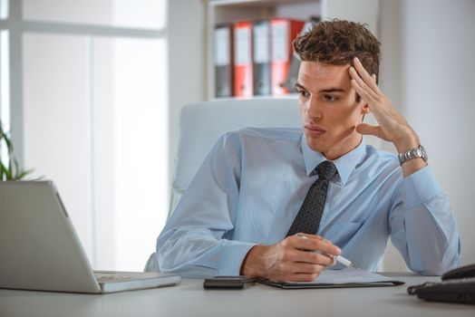 A serious young employee working in office with laptop and making notes, looking focused and concentrated.