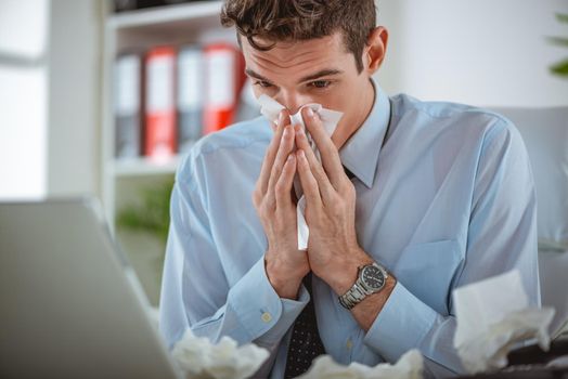 Young businessman blows his nose while working at his laptop in the office.
