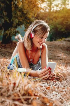 Cute smiling young woman lying down on the withered grass in early autumn sunny day, listening music.