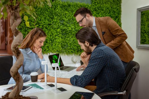 A team of a smart qualified skilled young engineers discussing about the project of alternative energy with miniatures of windmill turbines at the table.