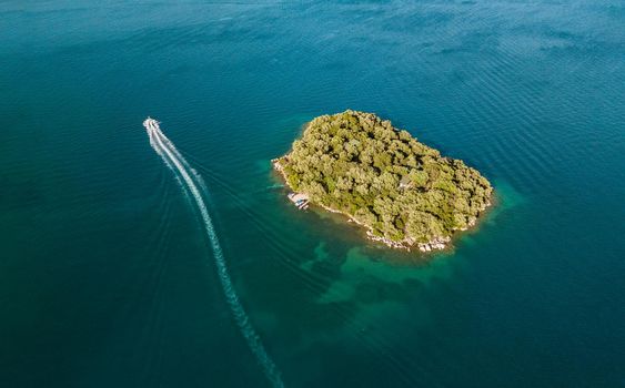 Drone shot of beautiful small island surrounded by crystal clear blue sea water and fast motorboat running away leaves a white trail in the waves of the sea.