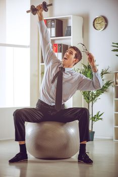 The young businessman sitting in the office on pilates ball and doing exercise with dumbbells.