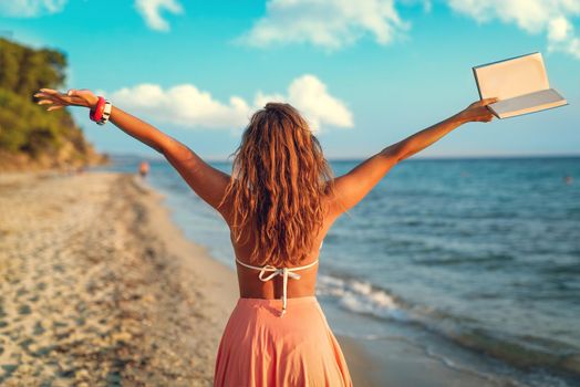 Rear view of a beautiful young woman enjoying on the beach with book in her open arms.