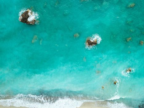 Drone shot of beautiful crystal clear turquoise and blue sea water, surrounding rocks and waves reaching sandy beach. 
