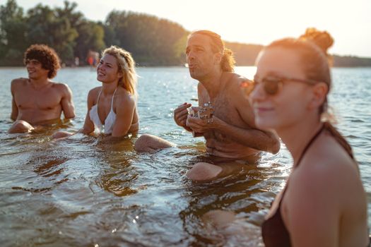 Happy young people having a great time together at the beach. They are sitting in the water and man is playing ukulele and singing. Sunset over water.