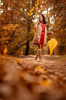 Happy young woman in red dress talking on smartphone walking in autumn sunny park, holding folded yellow umbrella.