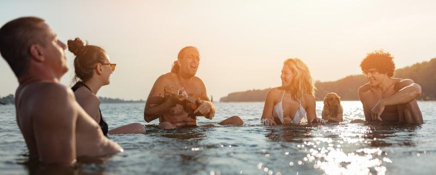 Happy young people having a great time together at the beach. They are sitting in the water and man is playing ukulele and singing. Sunset over water.