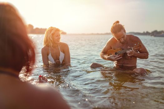 Happy young people having a great time together at the beach. They are sitting in the water and man is playing ukulele and singing. Sunset over water.