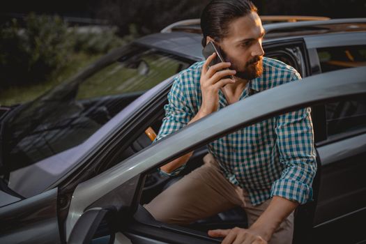 Young confident businessman holding smartphone in his hand and is talking, during is going to sit in the car.