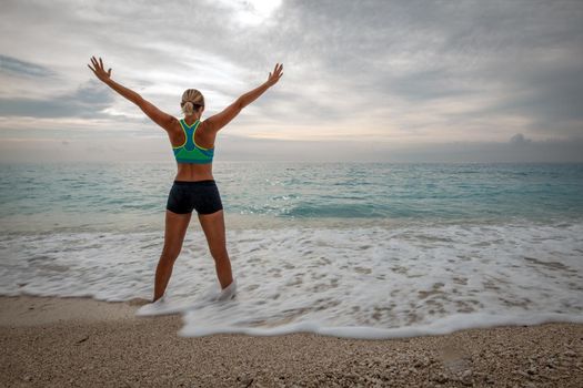 Beautiful young woman doing stretching exercise on the beach in the end of the day. Her arms are wide open. 