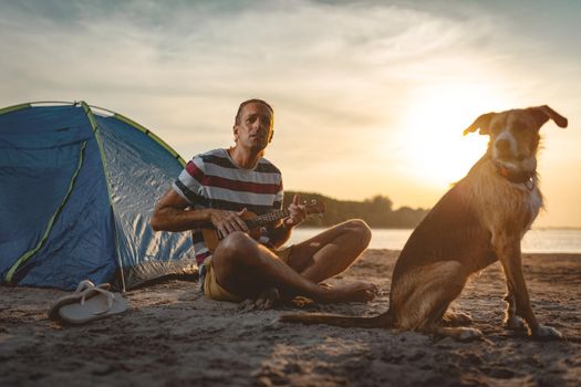 Happy young musician has a great time at the beach. He is sitting by the river with his dog, playing ukulele and singing. Sunset over water.