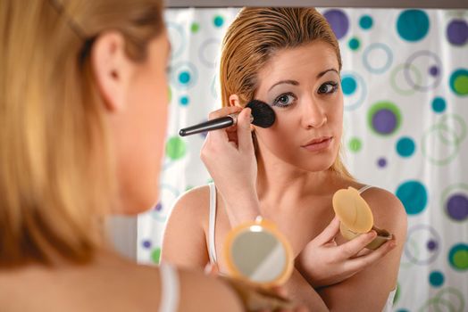 Young woman applying makeup on her face in front a mirror in the bathroom at home. 