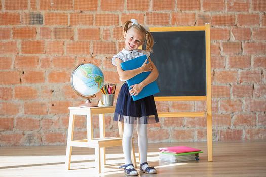 Adorable caucasian private school kindergarten girl smiling in front of blackboard with book in her hands, back to school concept