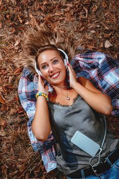 Top view of a cute girl lying down on the fall leaves and enjoying in listening music in the nature in early autumn sunny day.