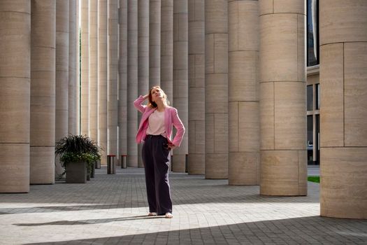 Young elegant beautiful blonde millennial with long hair in pink clothes stands and looks up in the colonnade of a modern building. Full height. Selective focus.