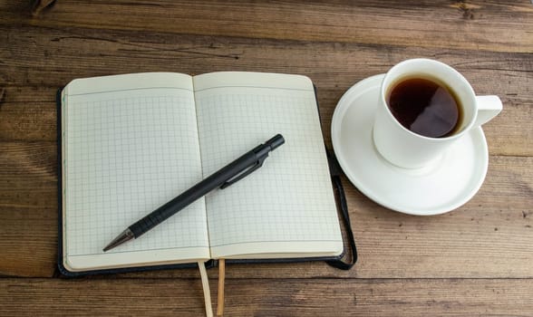 Coffee in a cup, notepad and pen on the table. Coffee in a cup with a saucer and a notebook with a pen on a wooden table.