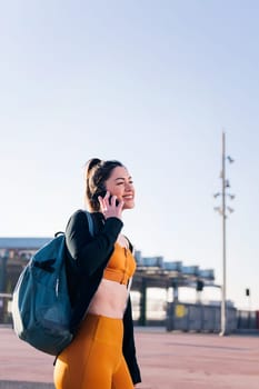 vertical portrait of a woman in sports clothes talking by mobile phone outdoors in the city street, concept of technology of communication and urban sport