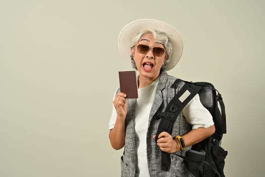 Smiling retired woman with backpack holding passport getting ready to travel on holidays on gray background.