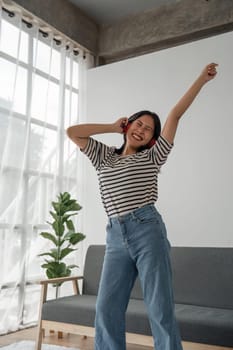 Attractive happy young Asian woman listening music with headphones from music application dance raise up hand near grey sofa couch stay at home rest relax spend free spare time in living room indoor.