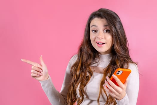 Happy woman with smartphone and shows by finger on the copy space, smiling at the camera and standing against a pink background.