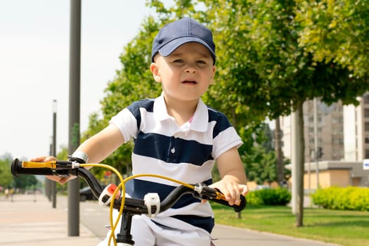 Sport concept. Boy on a bicycle close-up. A small beautiful, happy, Caucasian boy 4 years old in a cap and a striped T-shirt rides a bicycle along the bike path in the summer outdoors.