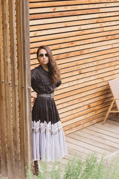 Lifestyle fashion portrait of young stylish hipster woman standing on the street, wearing trendy outfit, black dress. Wood background