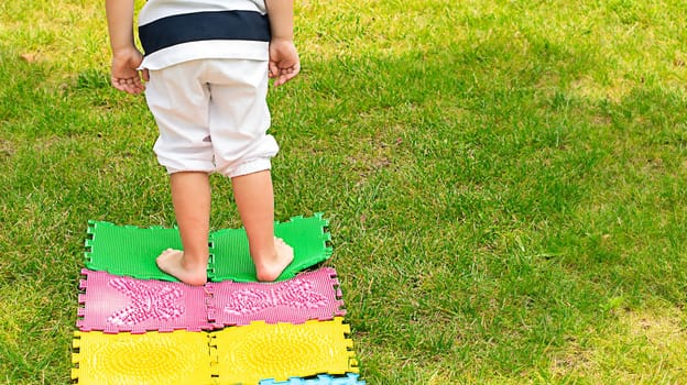 A little boy in white shorts stands on orthopedic multi-colored rugs. On the street in the park, on the green grass, he walks on rugs and massages his feet.