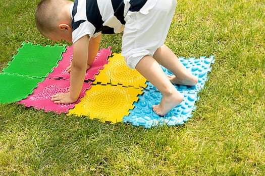 A little boy goes in for sports on massage orthopedic multi-colored rugs. A child in a striped T-shirt crawls merrily in the fresh air in the park, on green grass on massage mats. Stands in a plank position.