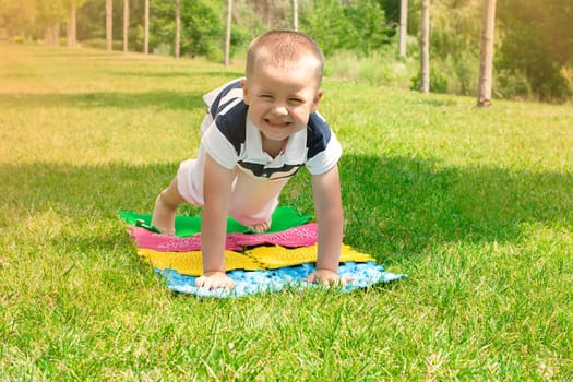 A little boy goes in for sports on massage orthopedic multi-colored rugs. A child in a striped T-shirt crawls merrily in the fresh air in the park, on green grass on massage mats. Stands in a plank position.
