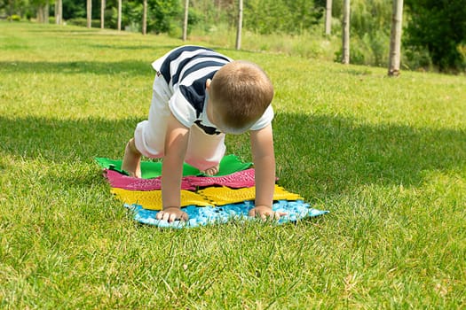 A little boy goes in for sports on massage orthopedic multi-colored rugs. A child in a striped T-shirt crawls merrily in the fresh air in the park, on green grass on massage mats. Stands in a plank position.