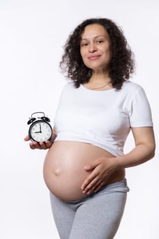 Charming pregnant woman gently strokes her belly, cutely smiles, shows alarm clock to camera with ticking biological time and countdown to the expected date of child birth. Waiting for baby. Pregnancy