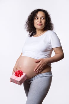 Beautiful curly multi ethnic pregnant woman, expectant mother, thoughtfully looking aside, touching her tummy, holding cute gift box near her naked belly, isolated on white. Happy Mother's Day concept