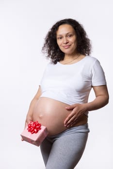 Attractive positive multi ethnic pregnant woman holding a gift box, touching and stroking her big belly in late pregnancy, smiling looking at camera, isolated over white studio background. Maternity