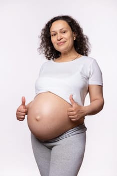 Multi ethnic curly beautiful pregnant woman gestures with thumbs up, expressing happiness and positive emotions, shows approval hand sign, smiles looking at camera, isolated on white studio background