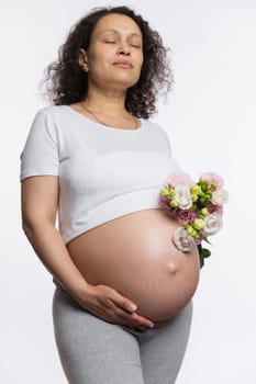 Delightful woman with a bouquet of spring flowers, caressing her big pregnant belly, posing with her eyes closed over isolated white background, enjoying happy emotions of her pregnancy and maternity