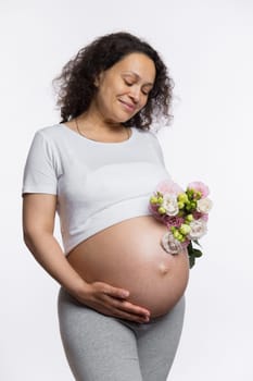 Delightful multi ethnic pregnant woman with a bunch of summer flowers, caressing her belly, smiling enjoying wonderful moments of her pregnancy and maternity, isolated white background. Expecting baby