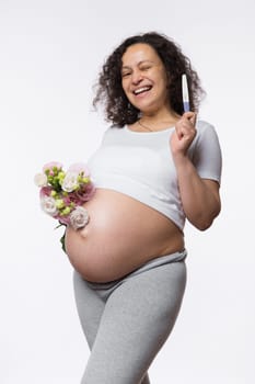 Smiling happy pregnant woman showing at camera a positive pregnancy test, posing with bunch of flowers, isolated over white background. Human fertility. Gynecology. Female health and maternity concept