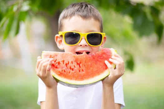 Cute boy in yellow sunglasses eating watermelon outdoors in summer. Healthy eating seasonal berries and fruits.
