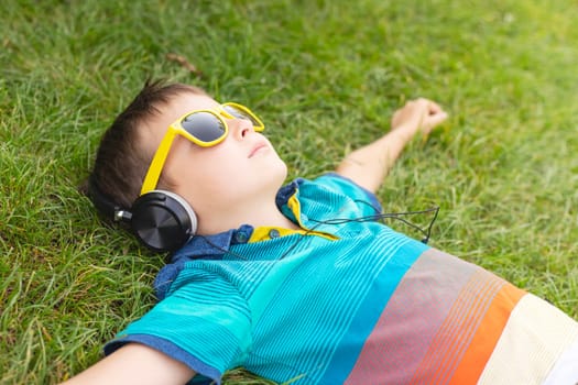 Happy boy in sunglasses lying on the grass and listening to music in headphones on a sunny day.