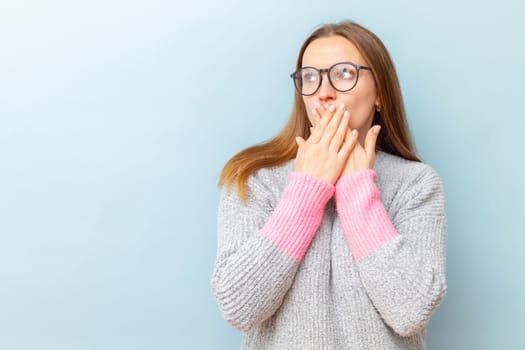 Shocked young woman in eyeglasses covering her mouth with her hands and looking at empty space for your text or object on blue background.